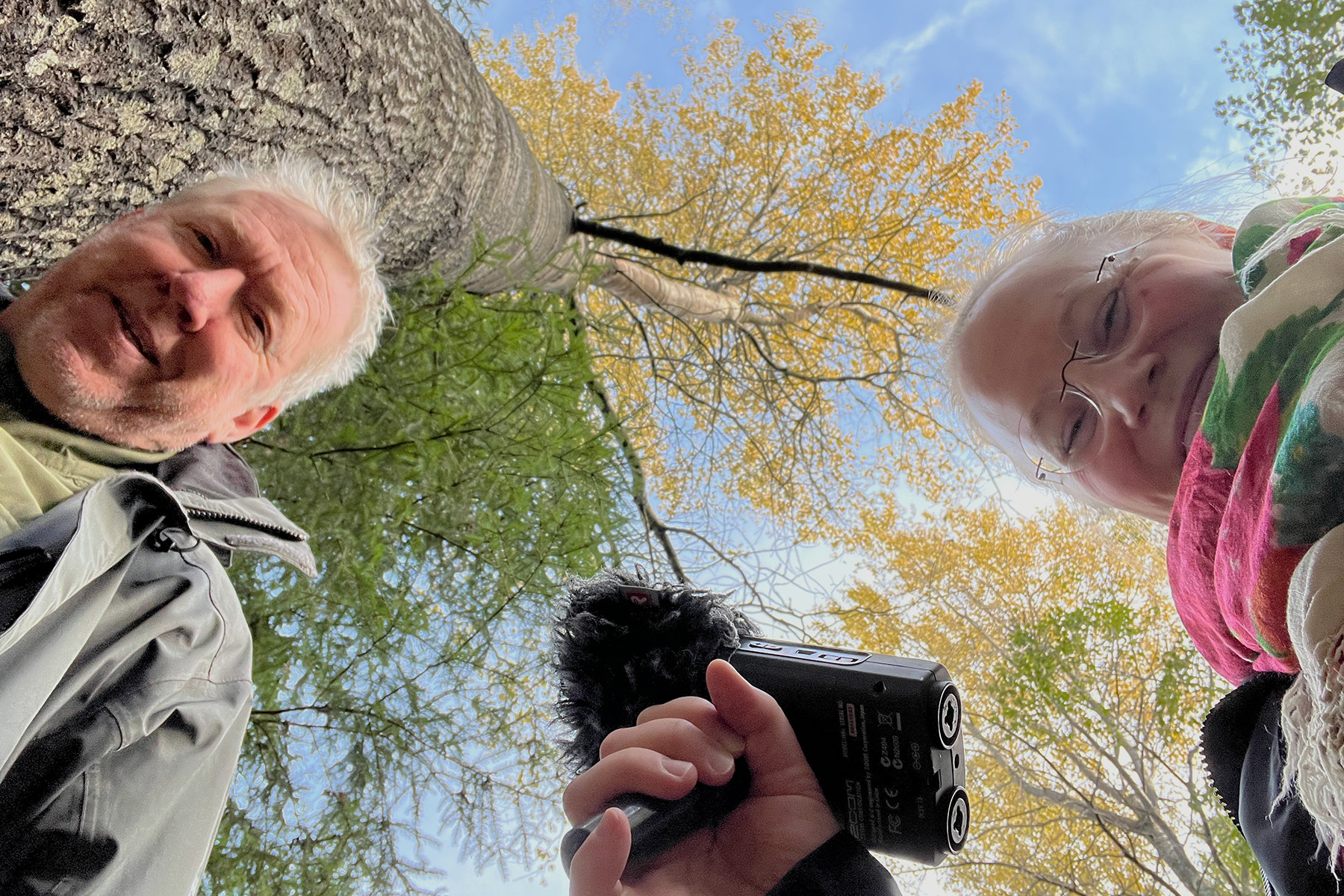 A woman stands on the right side looking into the camera and hold a voice recording device. A man stands on the left looking into the camera. The picture is taken from the frog perspective so that the woman and the man look downwards and tree tops are visible as the background.